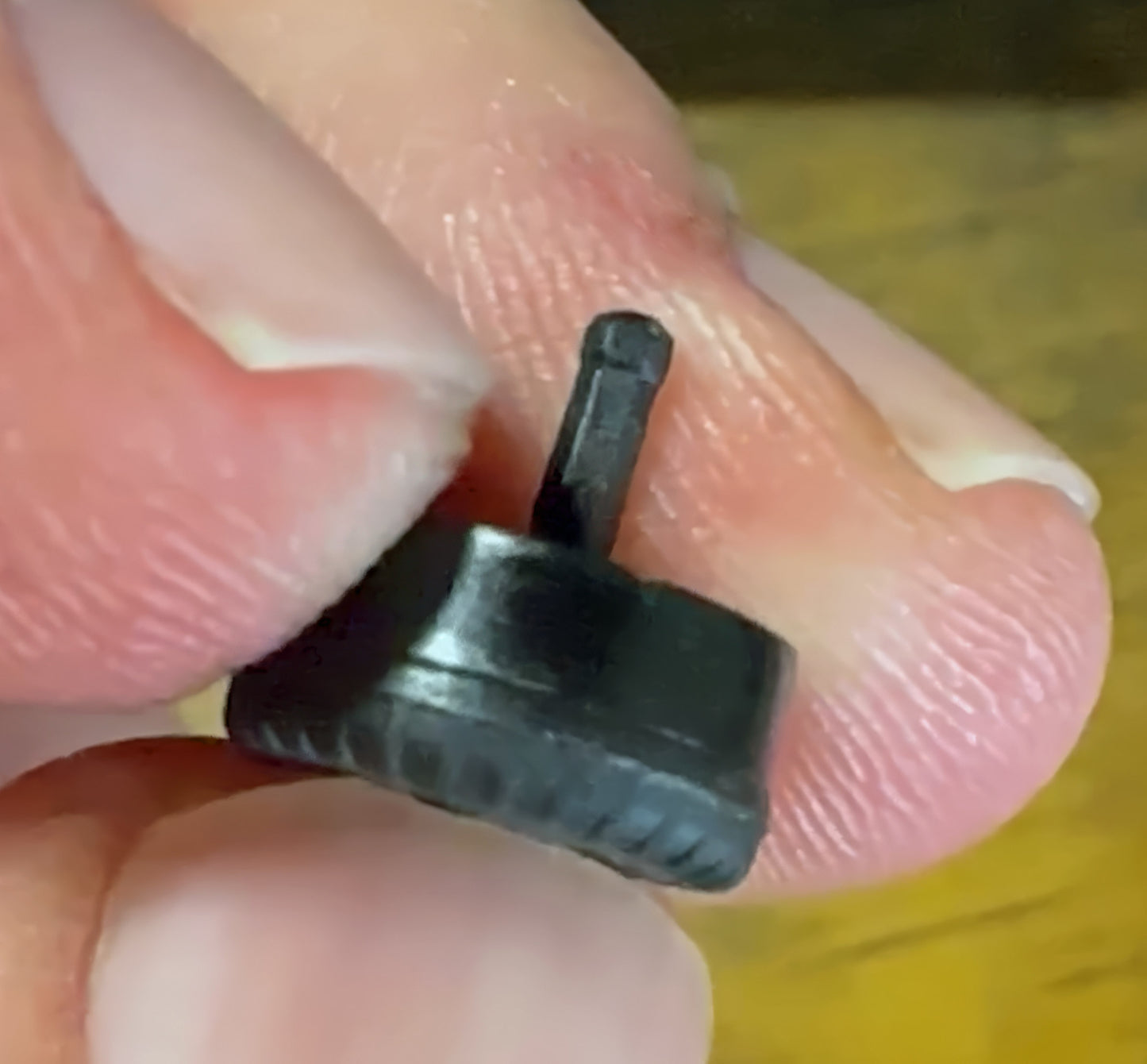 A close-up shows a hand holding a DermLite Replacement Power Button for DL5, resembling a small, black gear-like object with a protruding shaft. The fingers' texture is clear against the slightly blurred background.